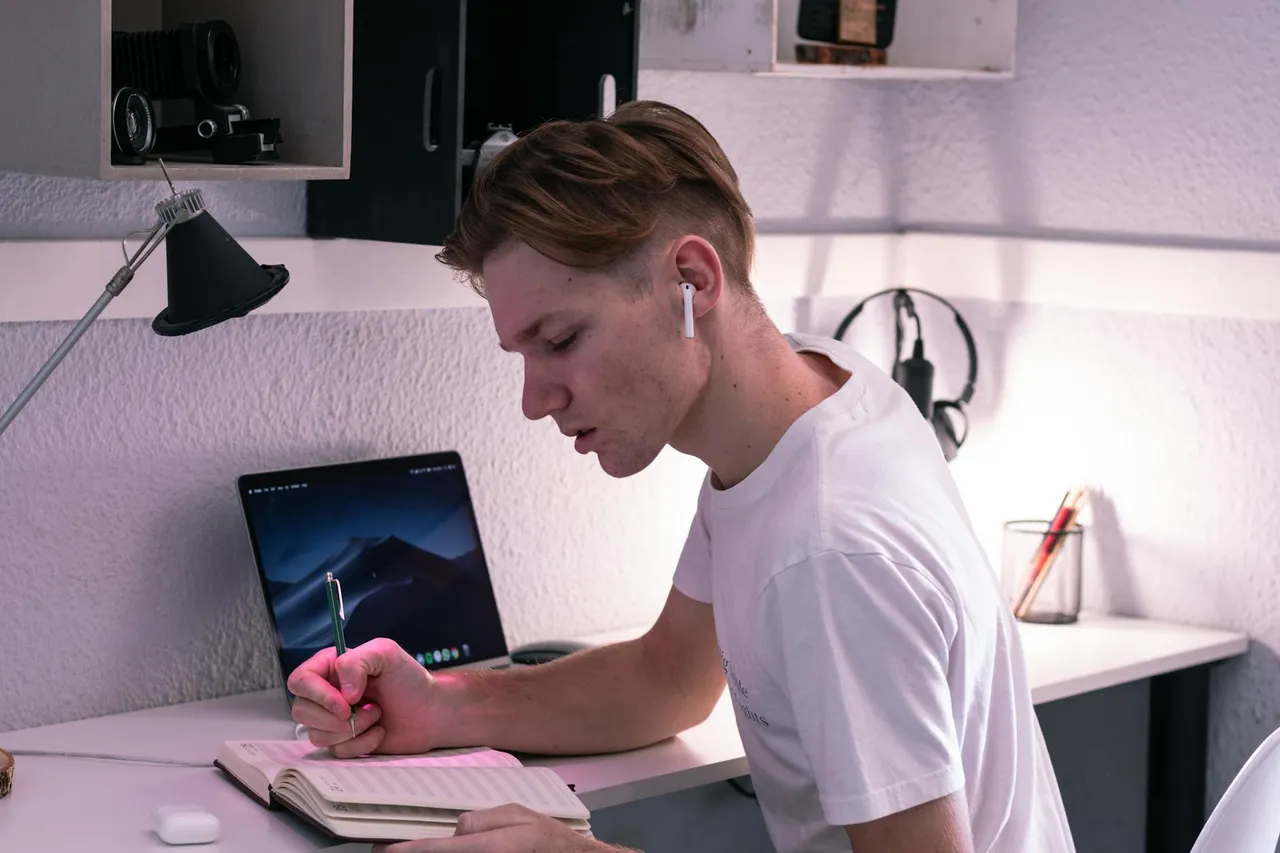 Side view of serious young man in TWS earbuds writing in notebook while studying on netbook at desk